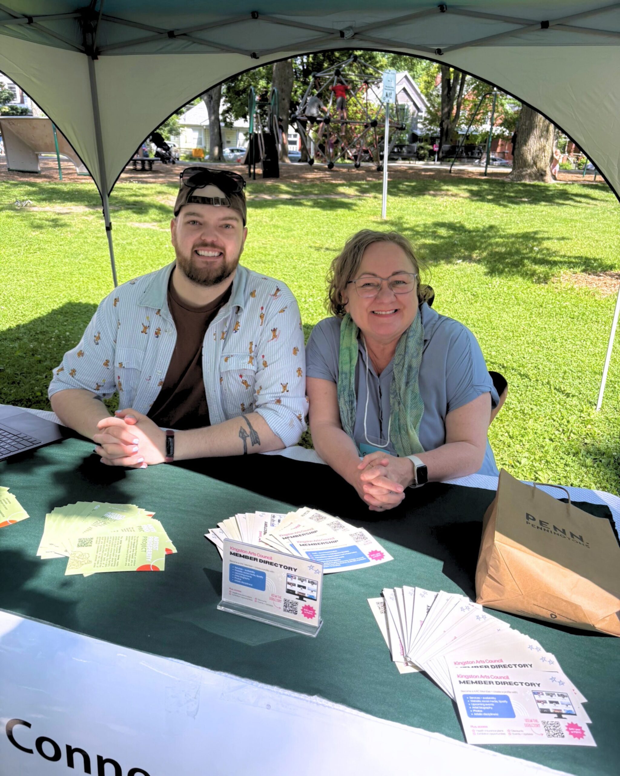 Staff member, B.C., and volunteer, Csilla, sit beside each other smiling at the Arts Council booth at the Skeleton Park Arts Festival