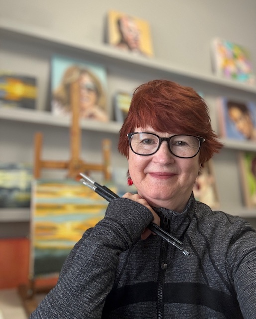 A photo of Ann Decker. Ann has bright red hair and is wearing black glasses and holding paint brushes. She smiles softly at the camera.