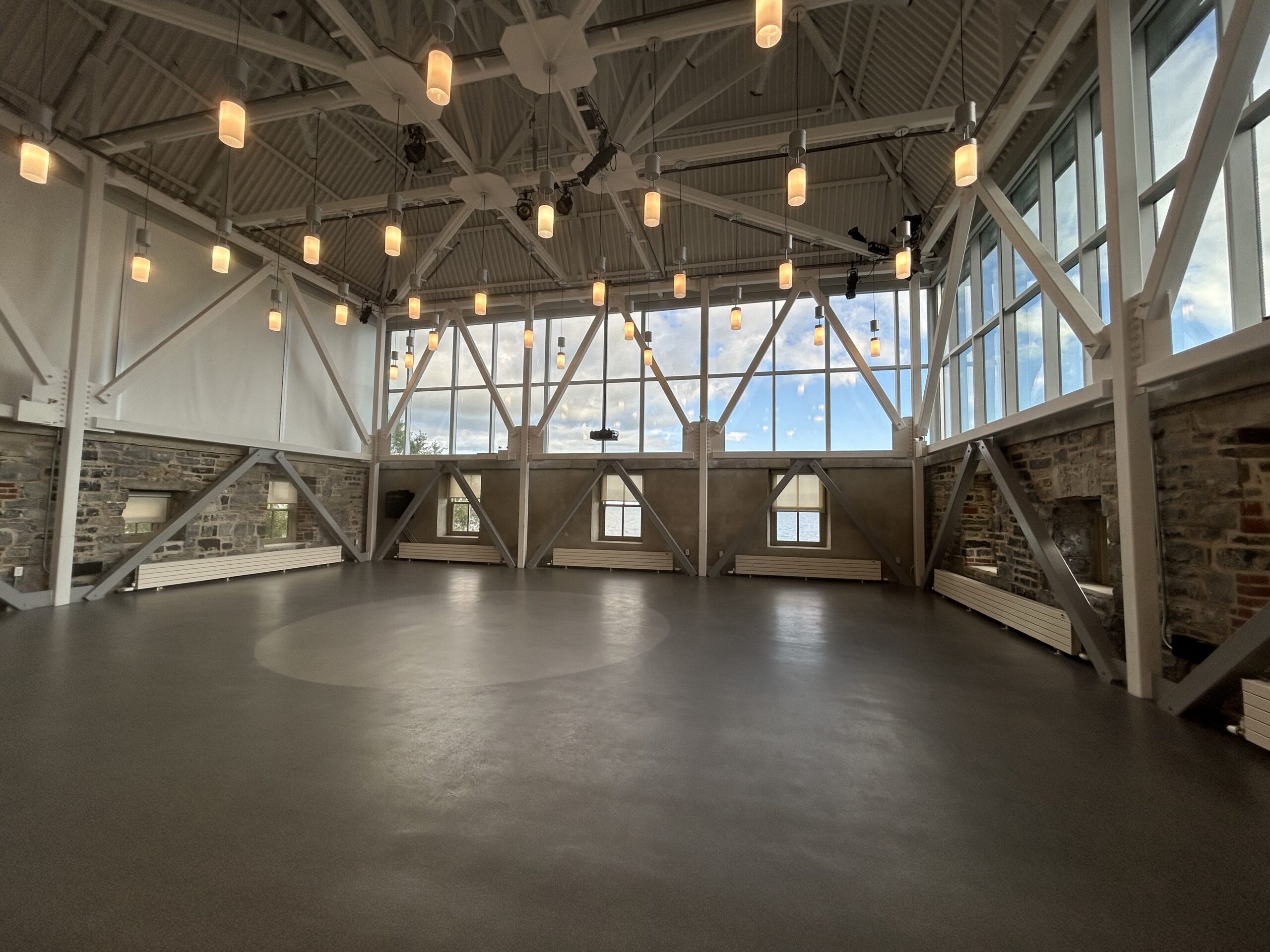 Malting Tower [large open event space with no furniture, bright with natural light coming through large surrounding windows, blinds closed on east side, high industrial vaulted ceilings with hanging light fixtures, support beams throughout the space with sections of limestone and concrete walls]