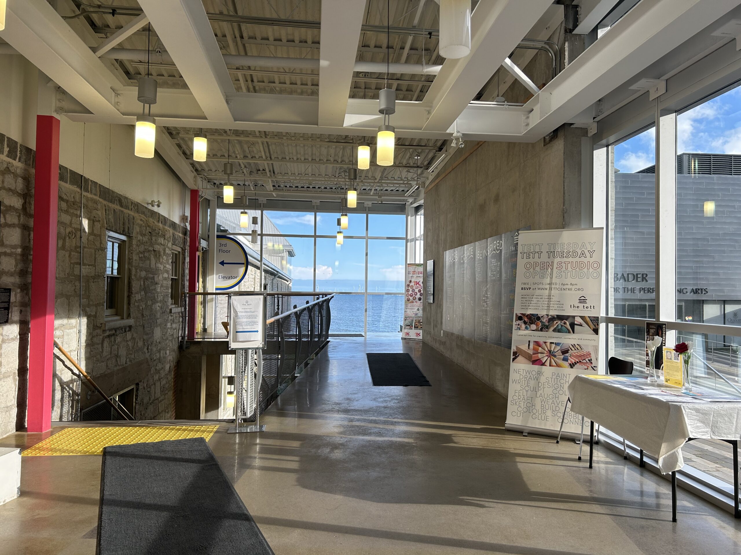 Entrance to the Tett Centre [open-space lobby area with bright windows on the west and south sides, staircase to the left heading downstairs with black carpet runner, information table along the west windows with standing Tett Tuesday banner beside, hallway leading towards the south windows and wrapping left to reach the door to the third floor and elevator]